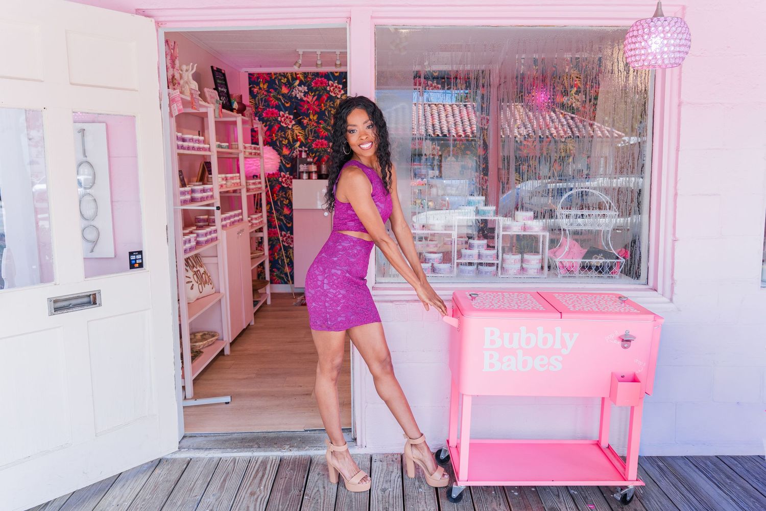 Woman in pink dress posing outside Bubbly Babes pink shop with matching pink cart on wooden deck.