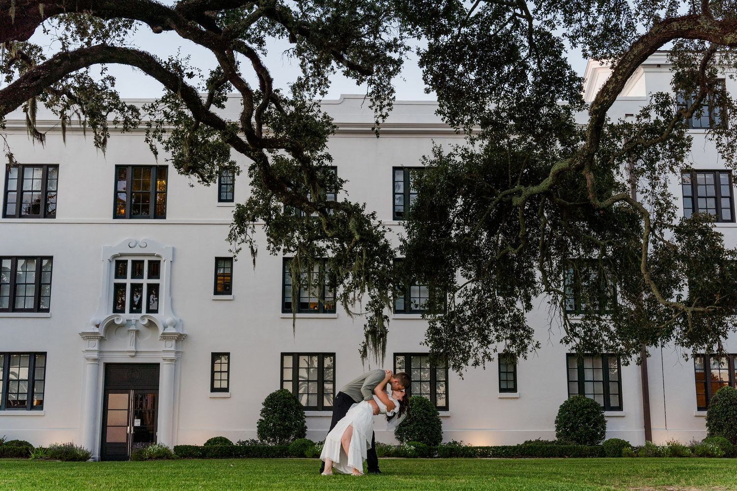 Historic white building with Spanish moss draped oak trees in foreground.
