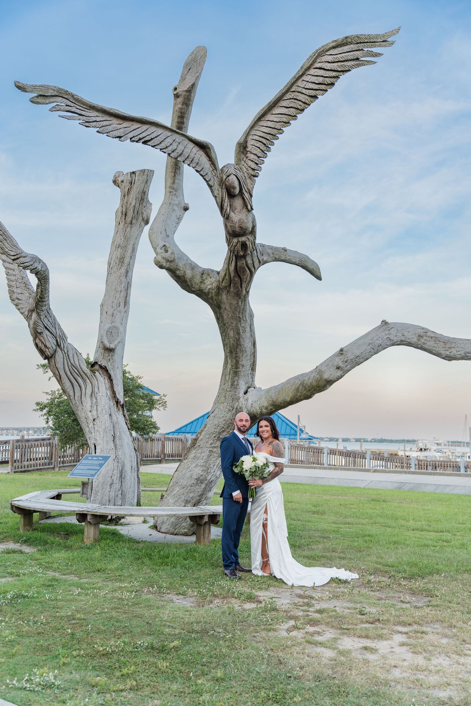 A wedding couple poses beneath unique driftwood sculptures on a grassy coastal area.