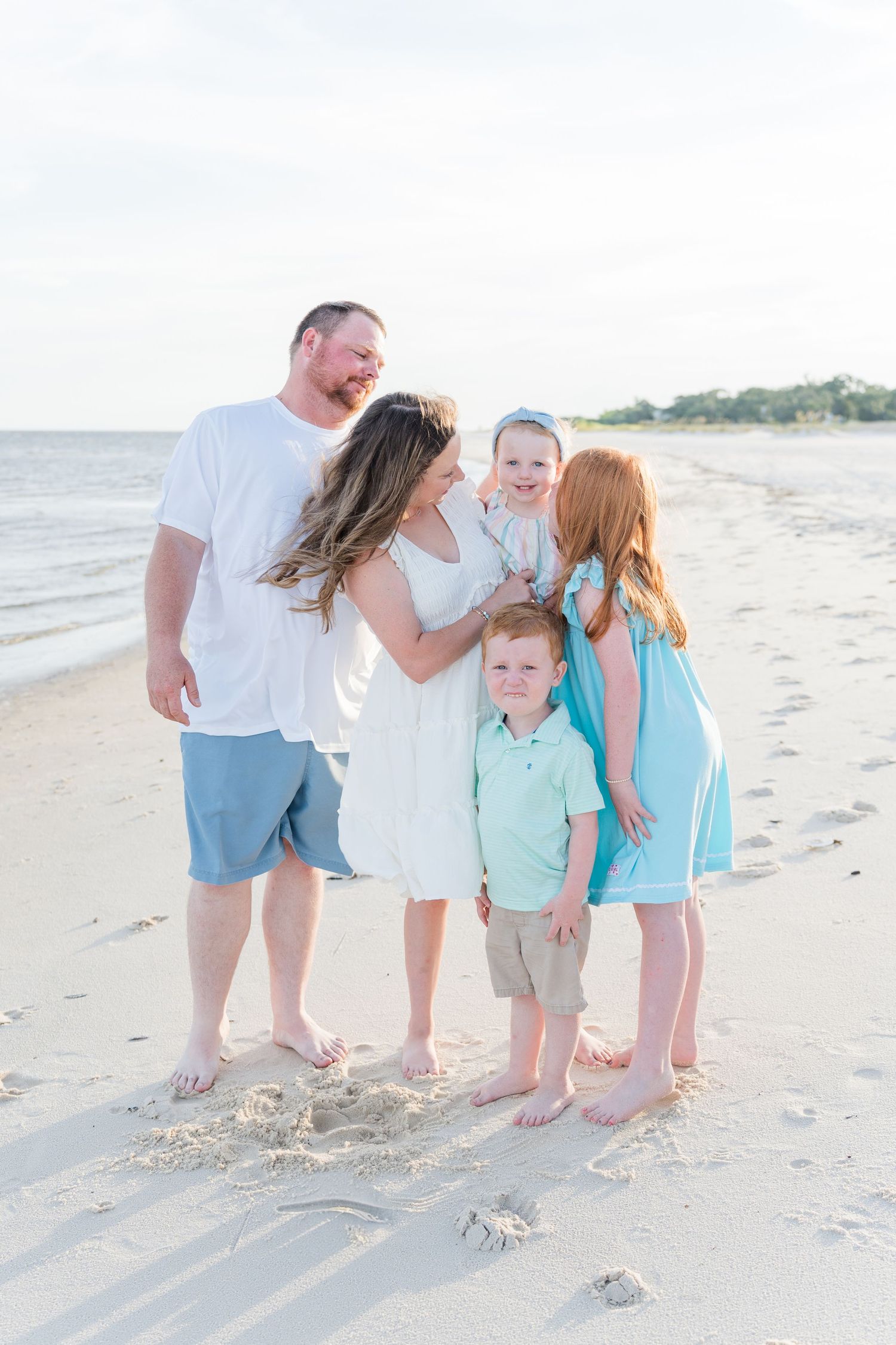 Family in coordinating beach outfits shares a tender moment on sandy shoreline.