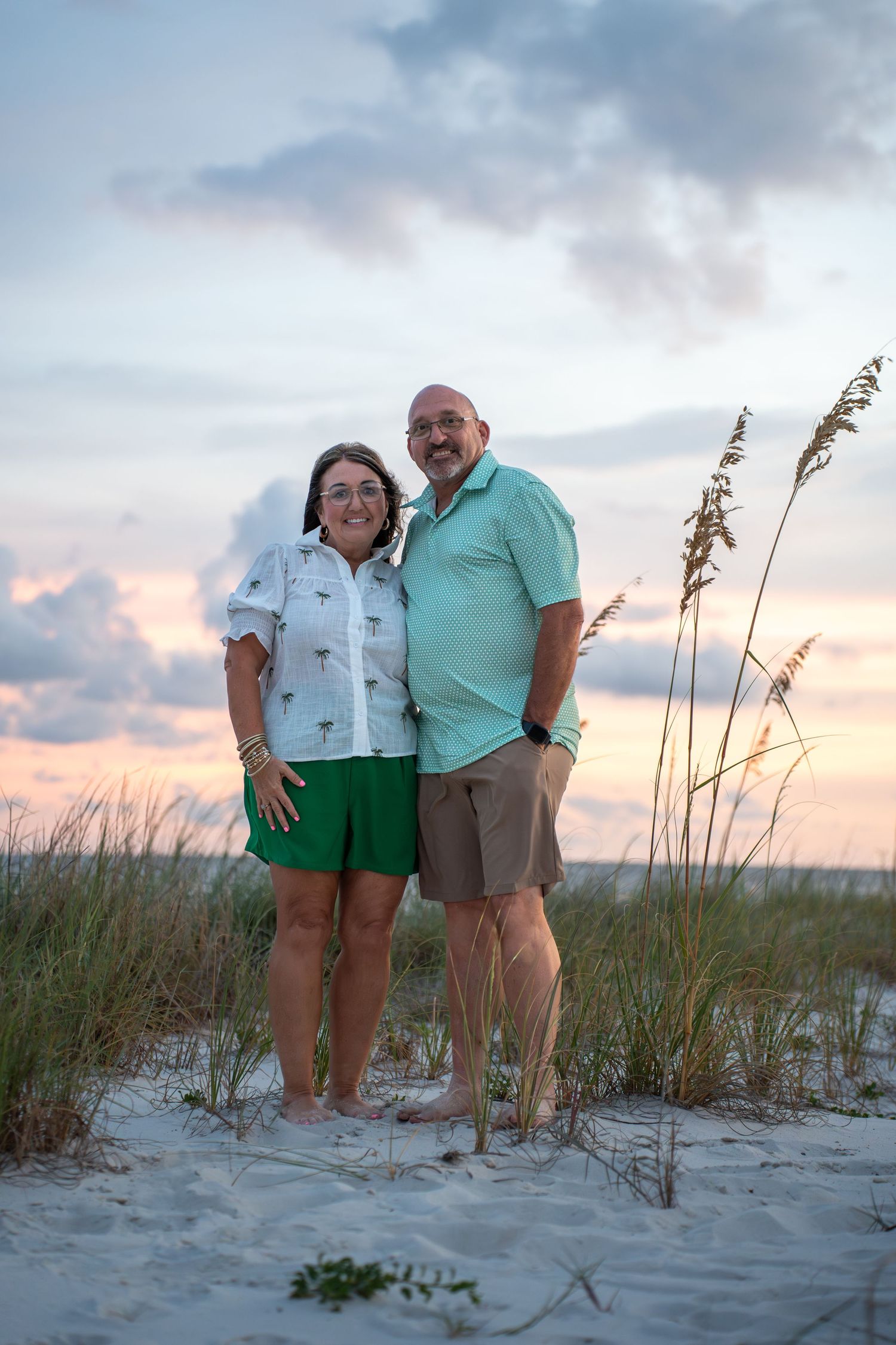 A couple poses together on a sandy beach with sea oats and a colorful sunset sky in the background.