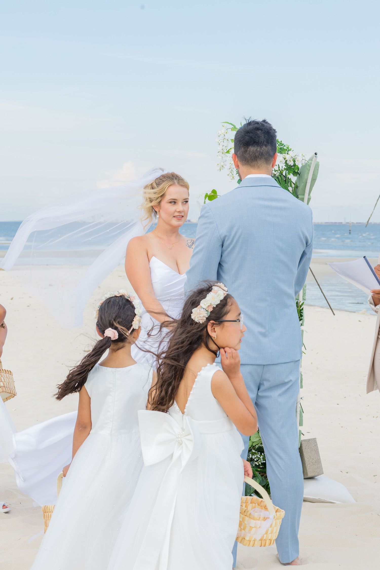 Bridesmaids in flowing white dresses gather on a sandy beach during a seaside wedding ceremony.