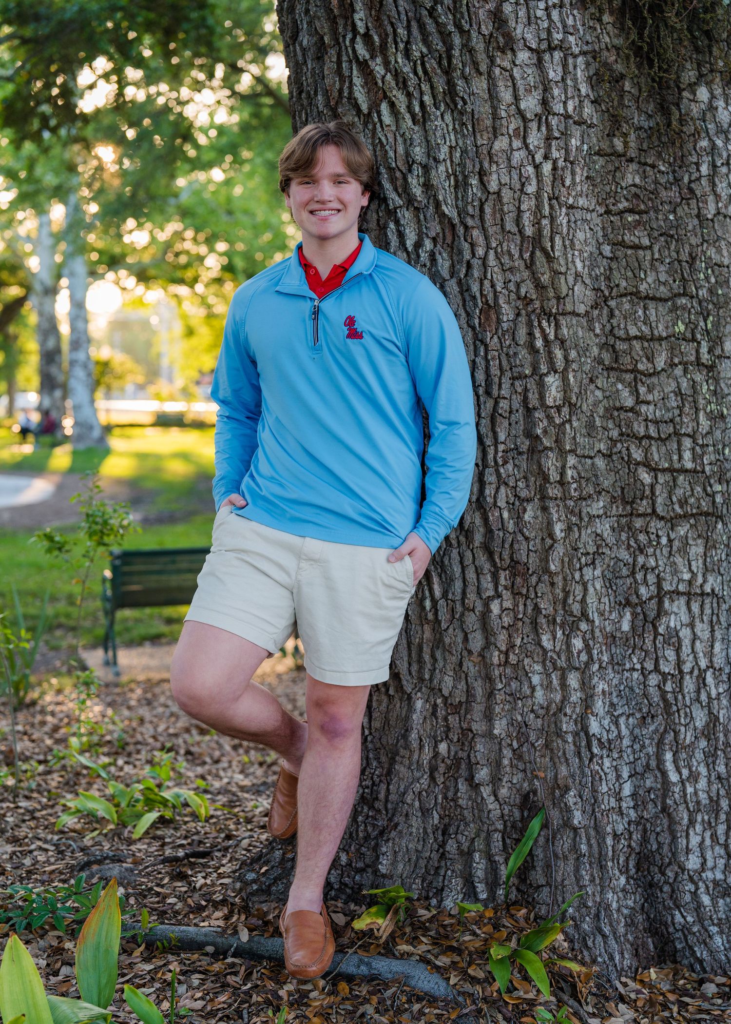 Outdoor senior portraits in blue polo and khaki shorts against large tree trunk.