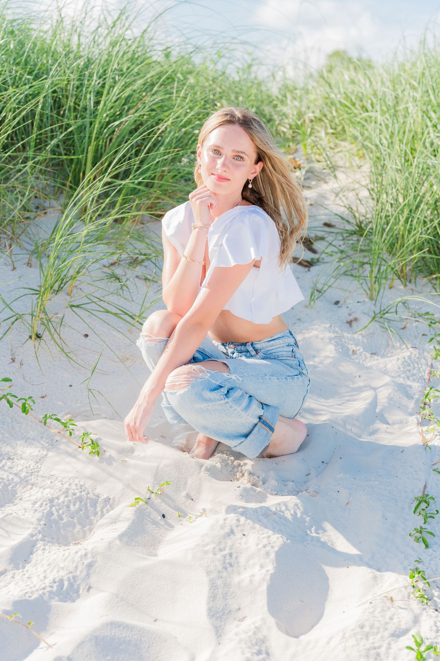 Woman in white top and jeans sits casually on sandy beach dunes with tall grass in background.