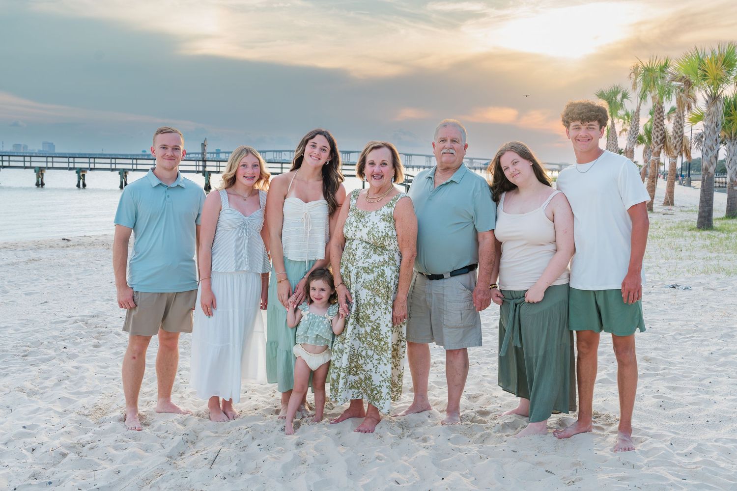 Family of eight posing on a sandy beach at sunset with palm trees and a pier in the background.