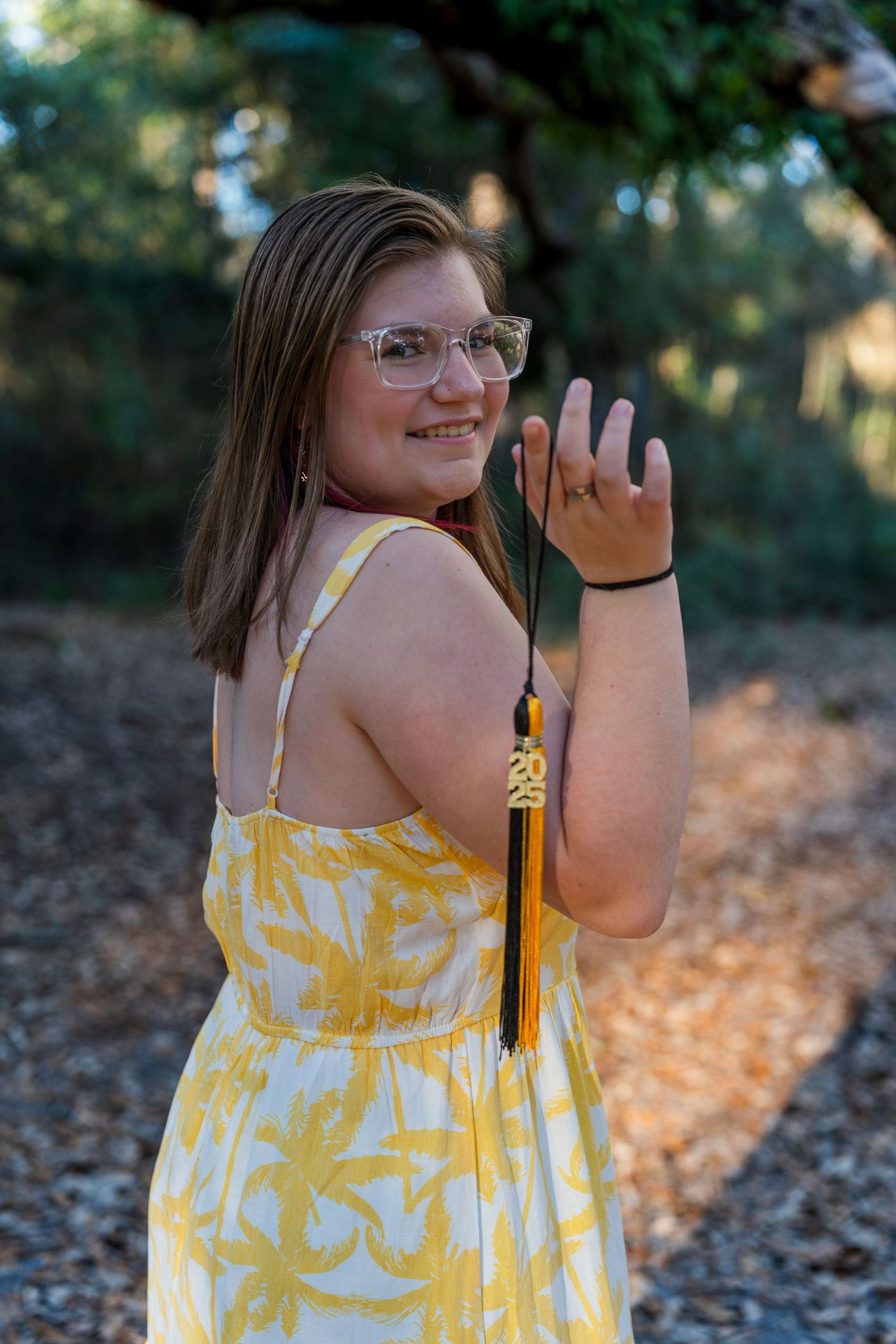 A person in a yellow dress poses with graduation tassel in a natural outdoor setting.