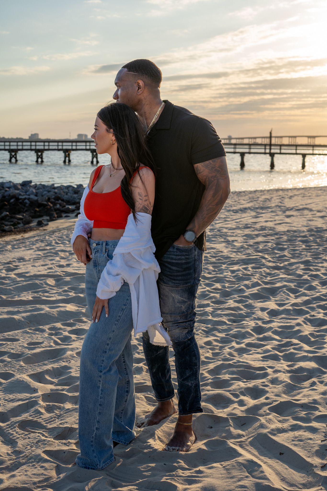 A couple embraces on a sandy beach with a pier visible in the sunset background.