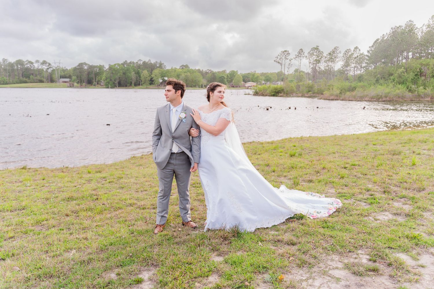Bride in flowing white gown and groom in gray suit pose by a serene lakeside on their wedding day.