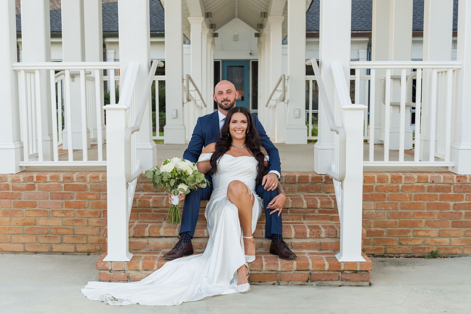 Newlyweds pose on brick steps of a white columned chapel entrance with wedding bouquet.