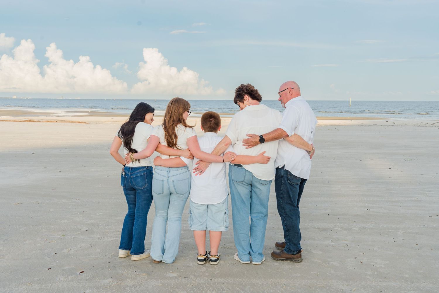 Family group hugging together on a sandy beach, viewed from behind with ocean and cloudy sky in background.