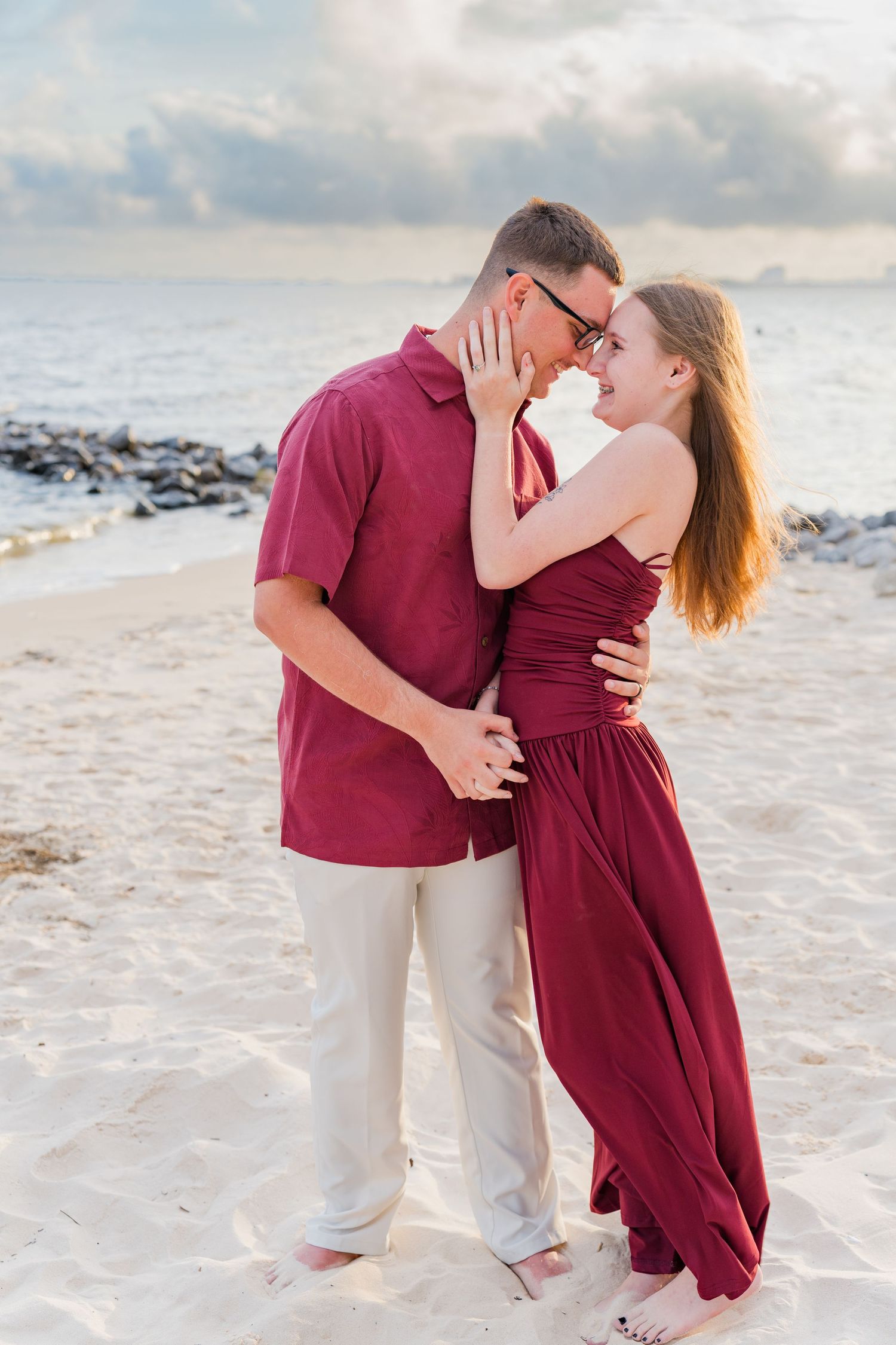 Couple in matching burgundy outfits embracing on a sandy beach, sharing a romantic moment near the ocean at dusk.