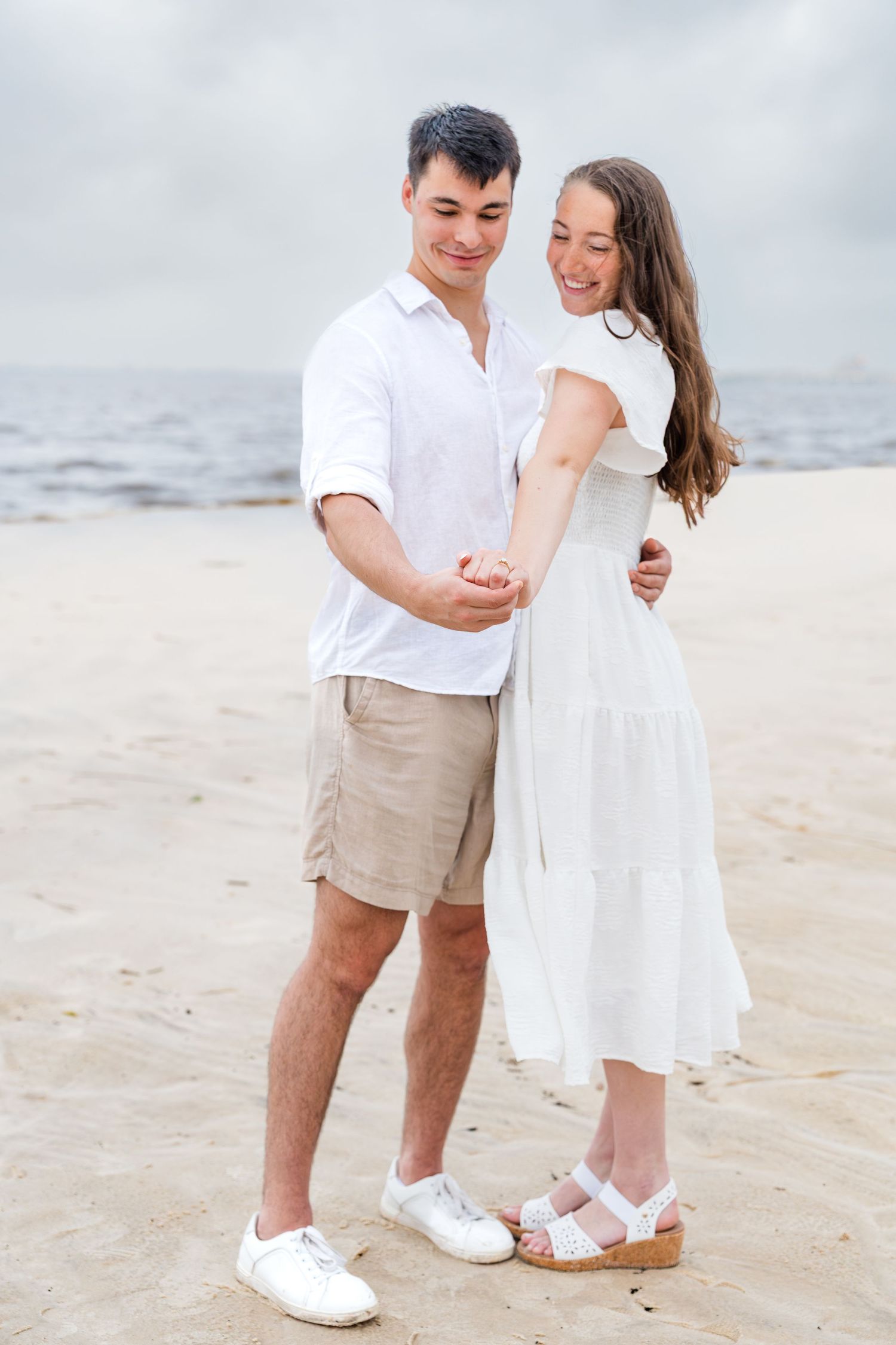 A happy couple in white outfits holding hands on a sandy beach with the ocean in the background.