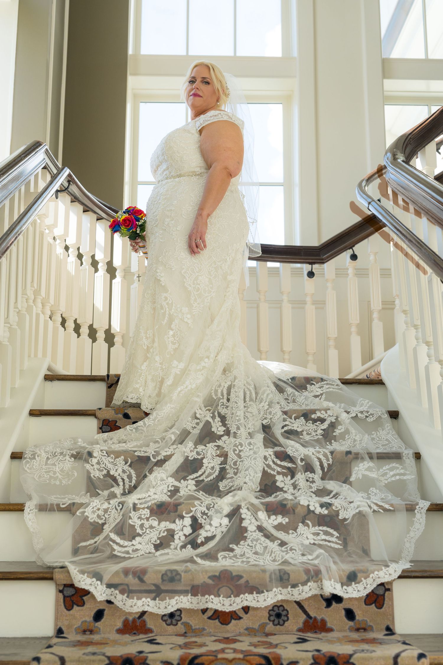 Bride in white lace gown with long train on grand staircase holding colorful bouquet.