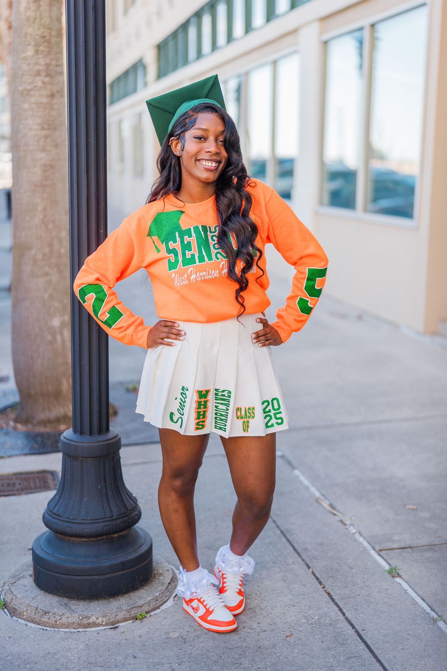 Graduate poses in orange and green university attire with white pleated skirt near lamp post.
