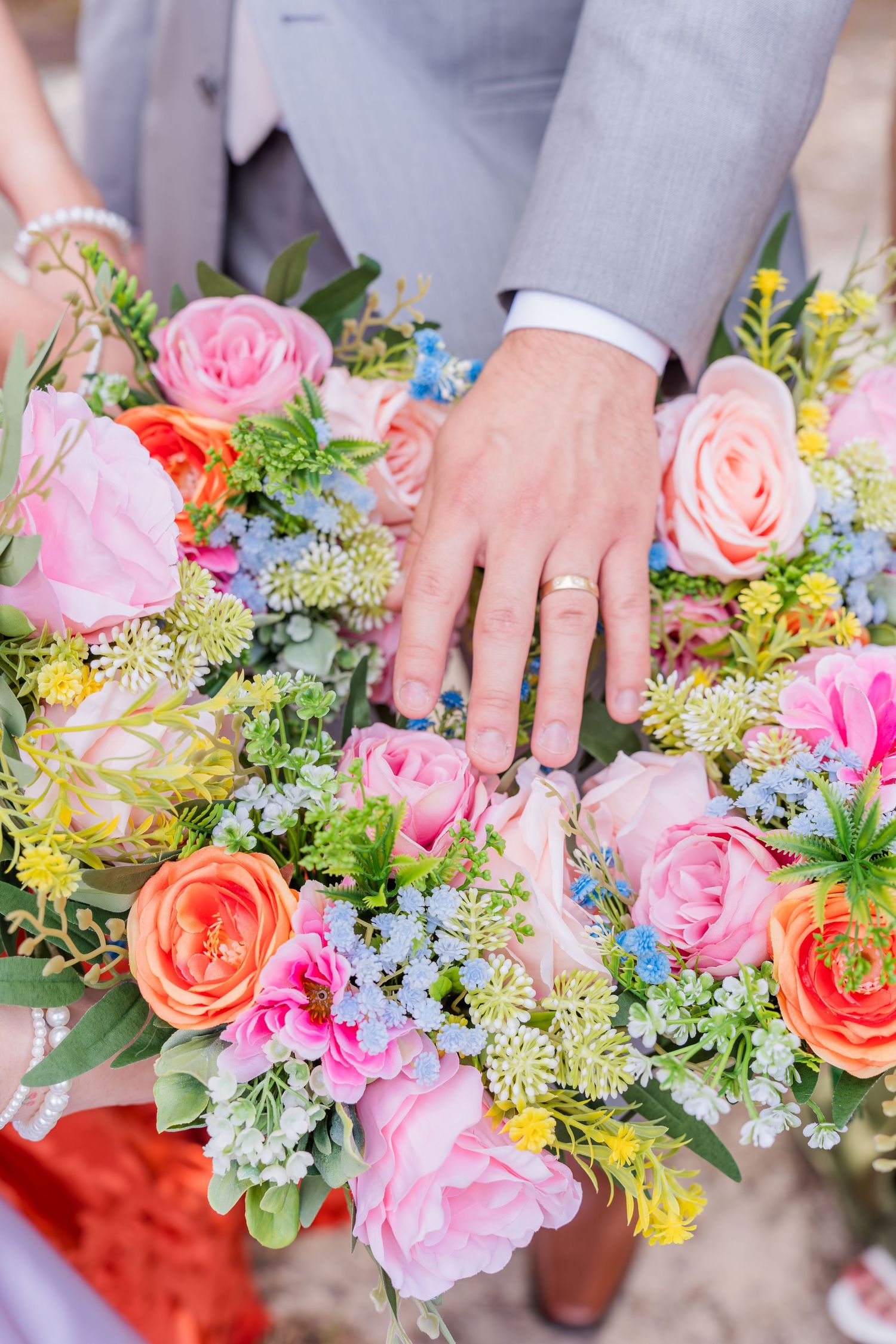 Newlyweds hands with gold wedding rings resting on a colorful bridal bouquet of pink, orange roses and wildflowers.
