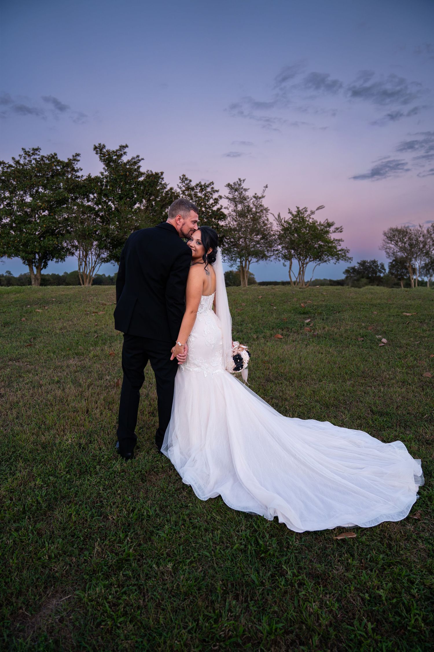 A couple in wedding attire stands on a grassy hillside at dusk with a flowing white dress trailing behind them.
