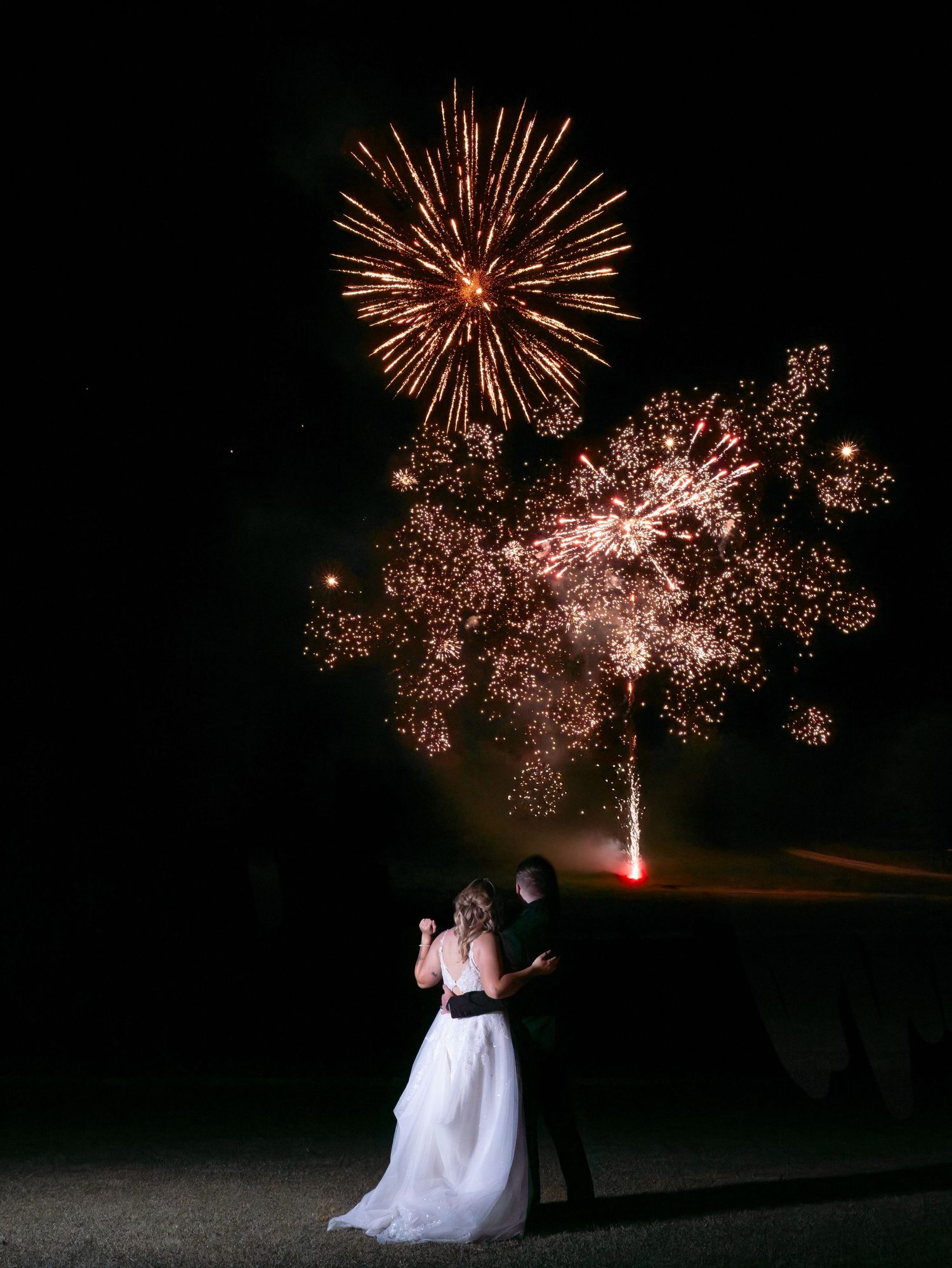 Romantic wedding celebration with fireworks display lighting up the night sky behind couple in formal attire.