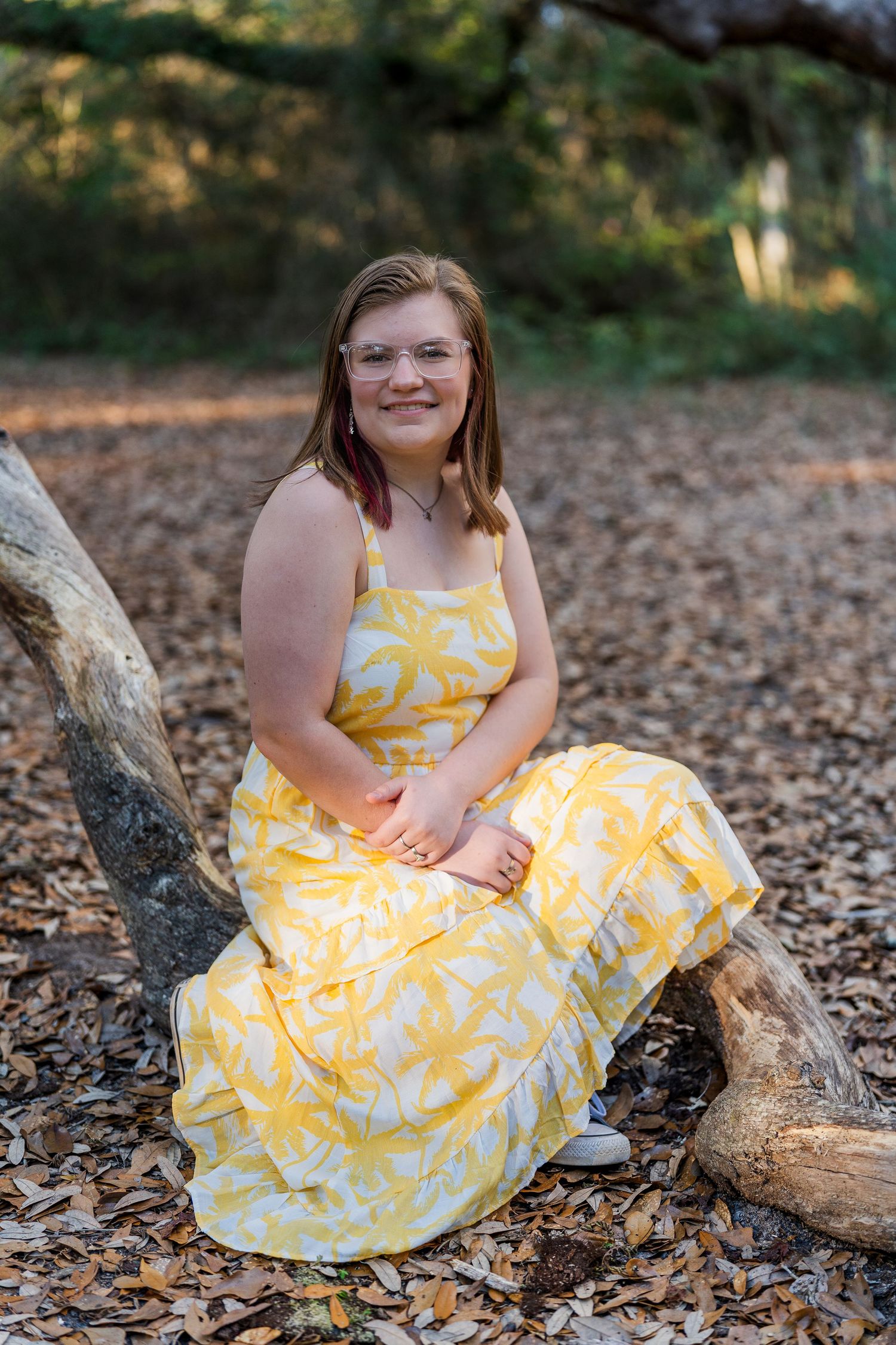 A person in a yellow floral sundress sits on a fallen tree trunk in a wooded area.