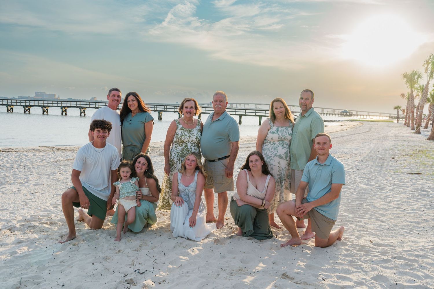 Family group photo on a sandy beach at sunset, wearing coordinated teal and green outfits near a pier.