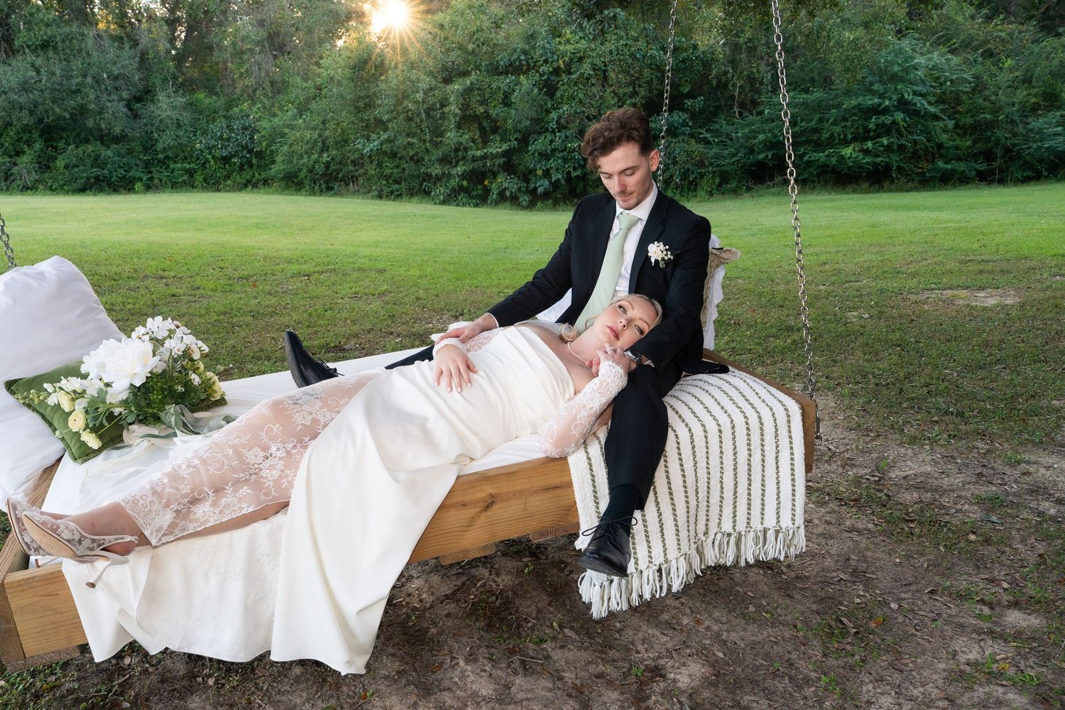 Bride in a white wedding gown rests her head on the groom's lap while they sit on a wooden swing beside a bouquet at sunset.