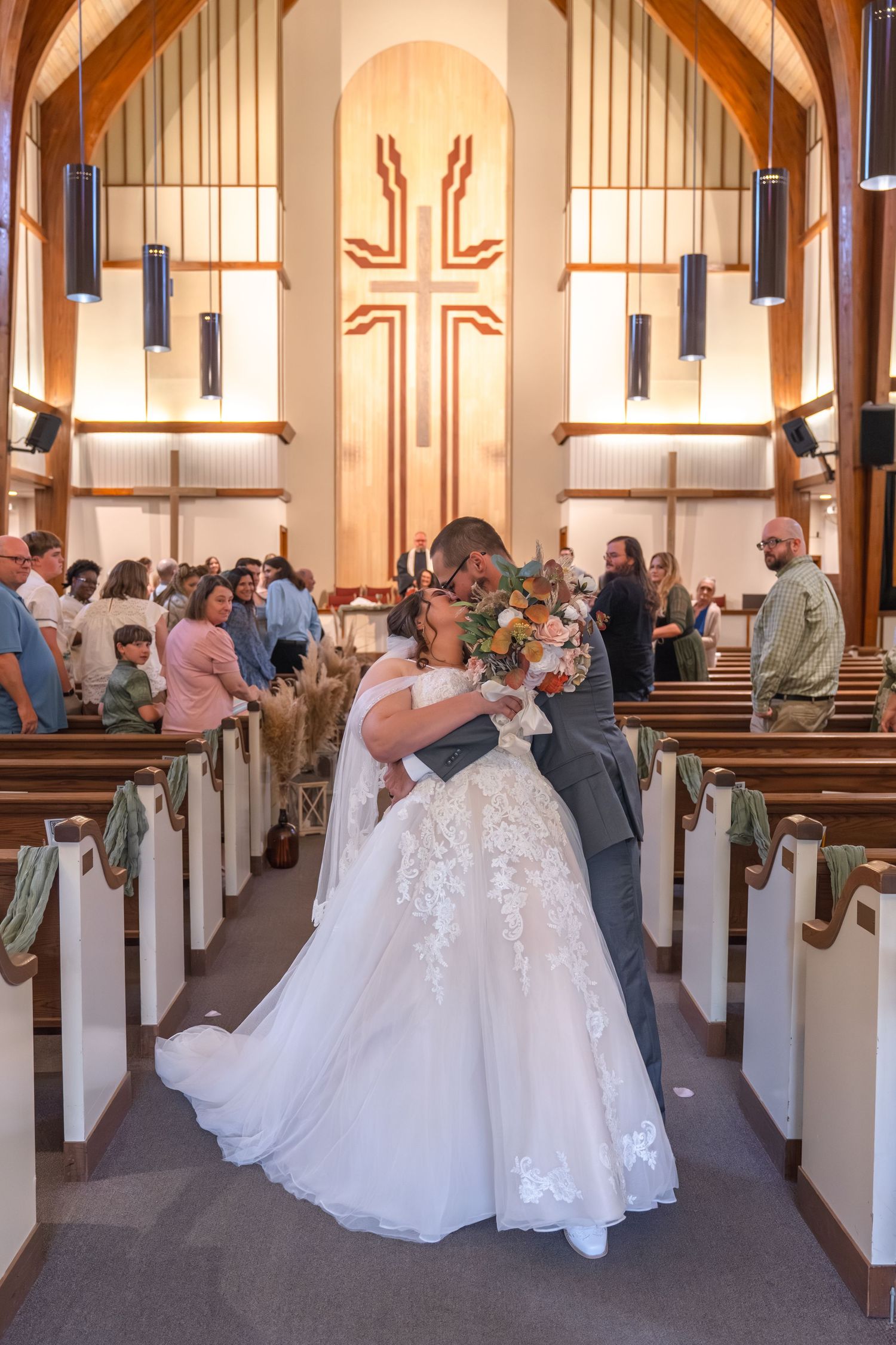 A couple embraces during their wedding ceremony in a church sanctuary with wooden pews and a large cross on the wall.