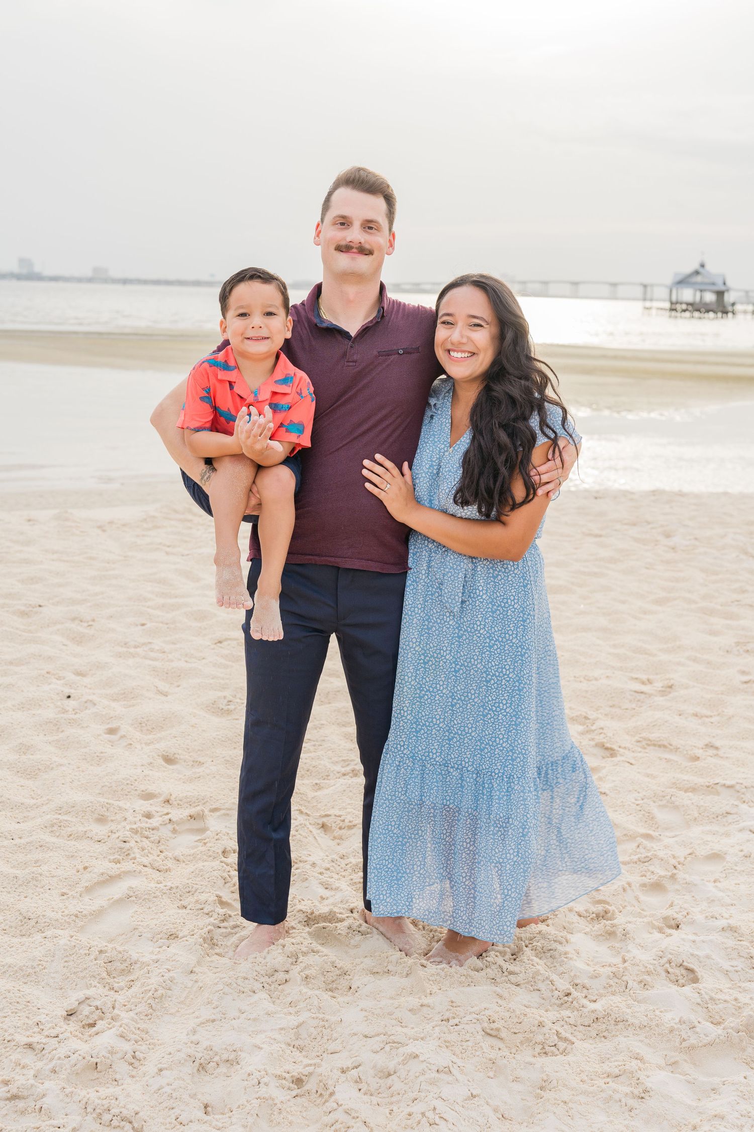 A family poses together on a sunny beach with the ocean visible in the background.
