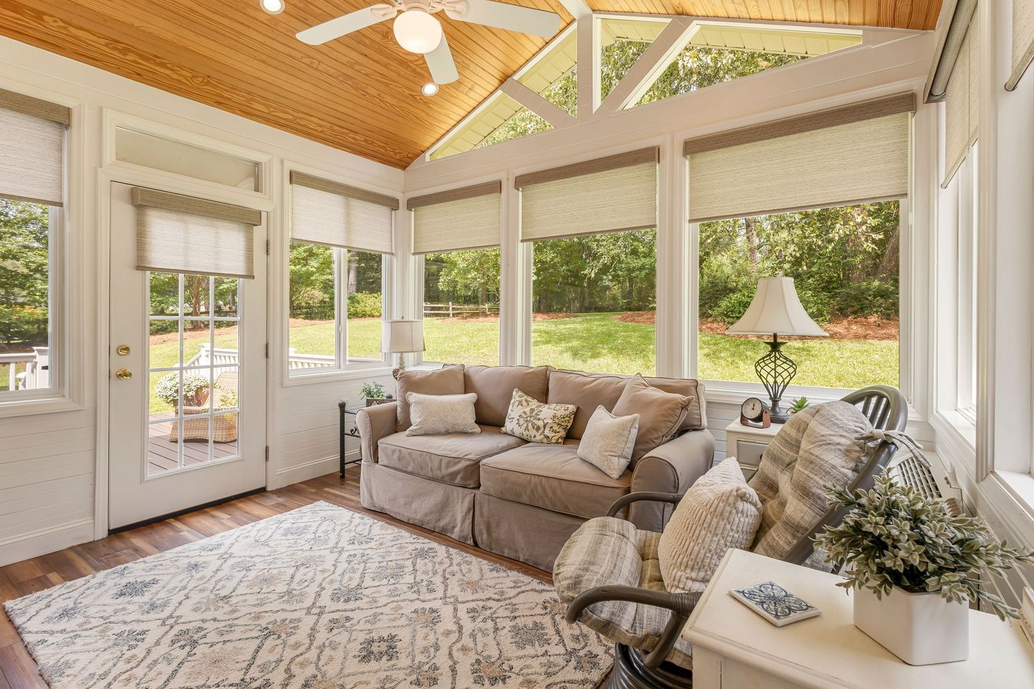 Bright sunroom with vaulted wood ceiling, large windows, gray sofa, patterned rug, and lush garden views.