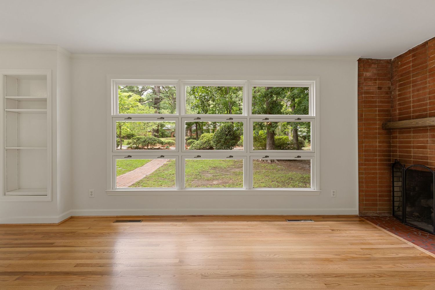 Bright living room with hardwood floors, large window overlooking garden, built-in shelves, and brick fireplace.