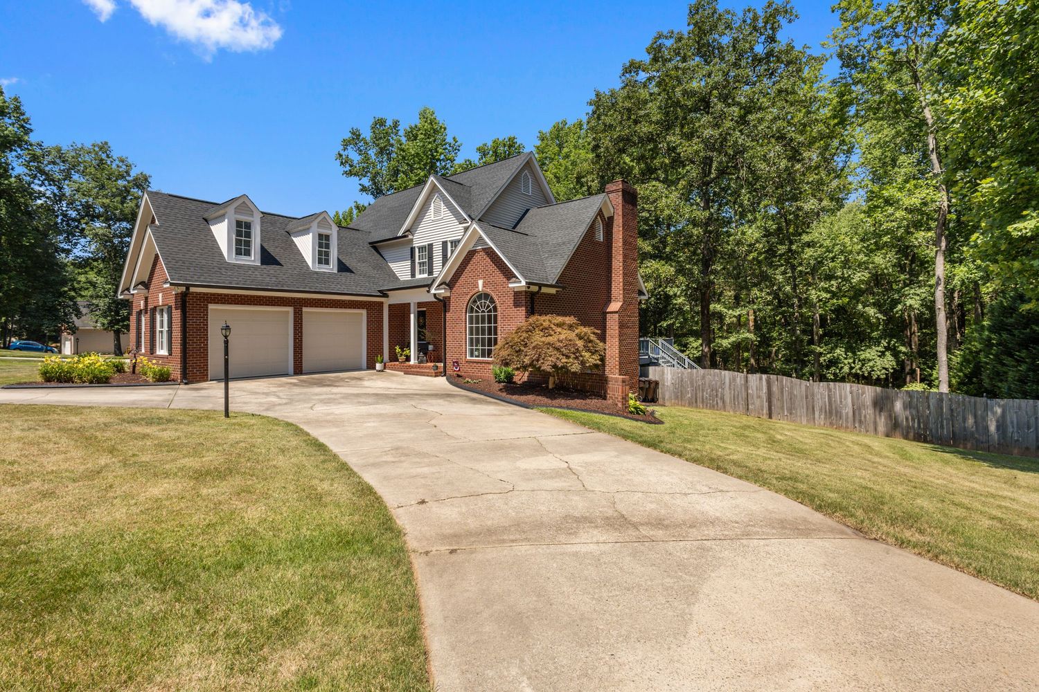 Brick two-story home with attached two-car garage, curved driveway, manicured lawn, and mature trees in a suburban setting.