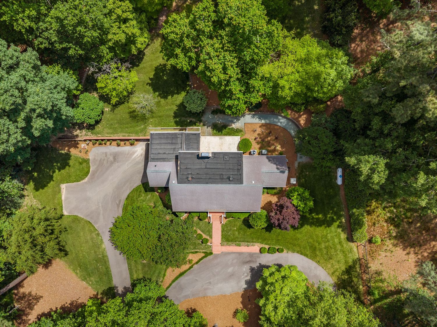 Aerial view of a suburban home surrounded by lush green trees, featuring a gray roof, brick driveway, and circular path.