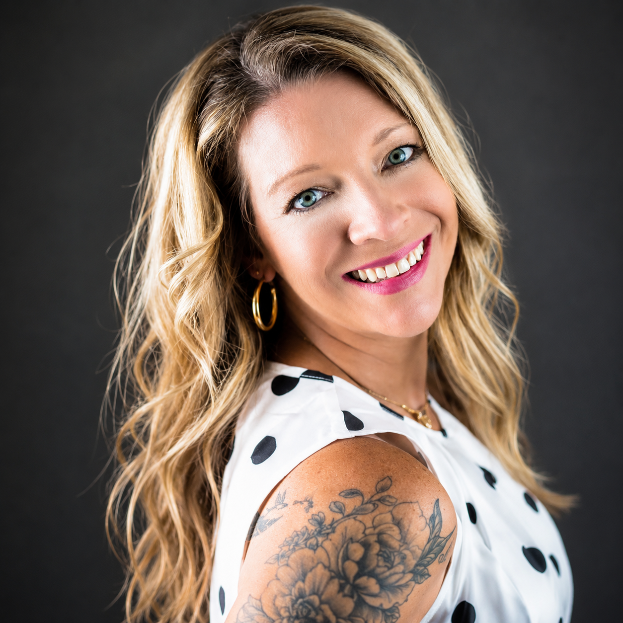 Smiling blonde woman with blue eyes, hoop earrings, floral shoulder tattoo, wearing a white polka dot top.