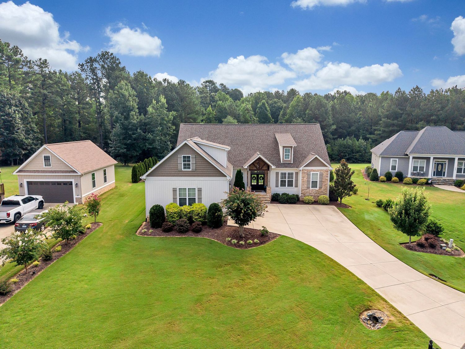 Aerial view of a large craftsman-style home with lush green lawn, landscaped beds, and wooded backdrop on a sunny day.