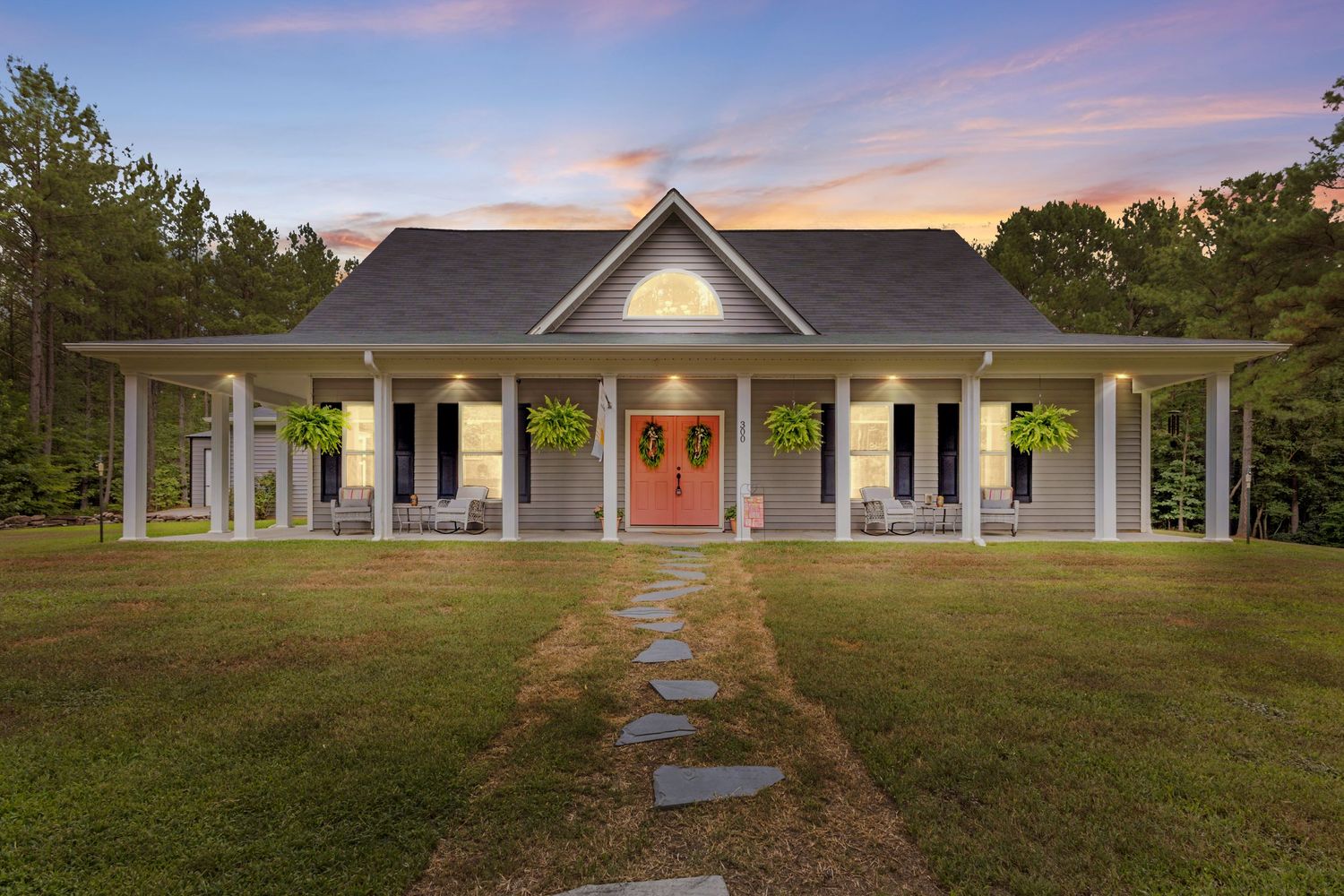 Charming white farmhouse with wrap-around porch, red double doors, and stone path at dusk surrounded by lush trees.