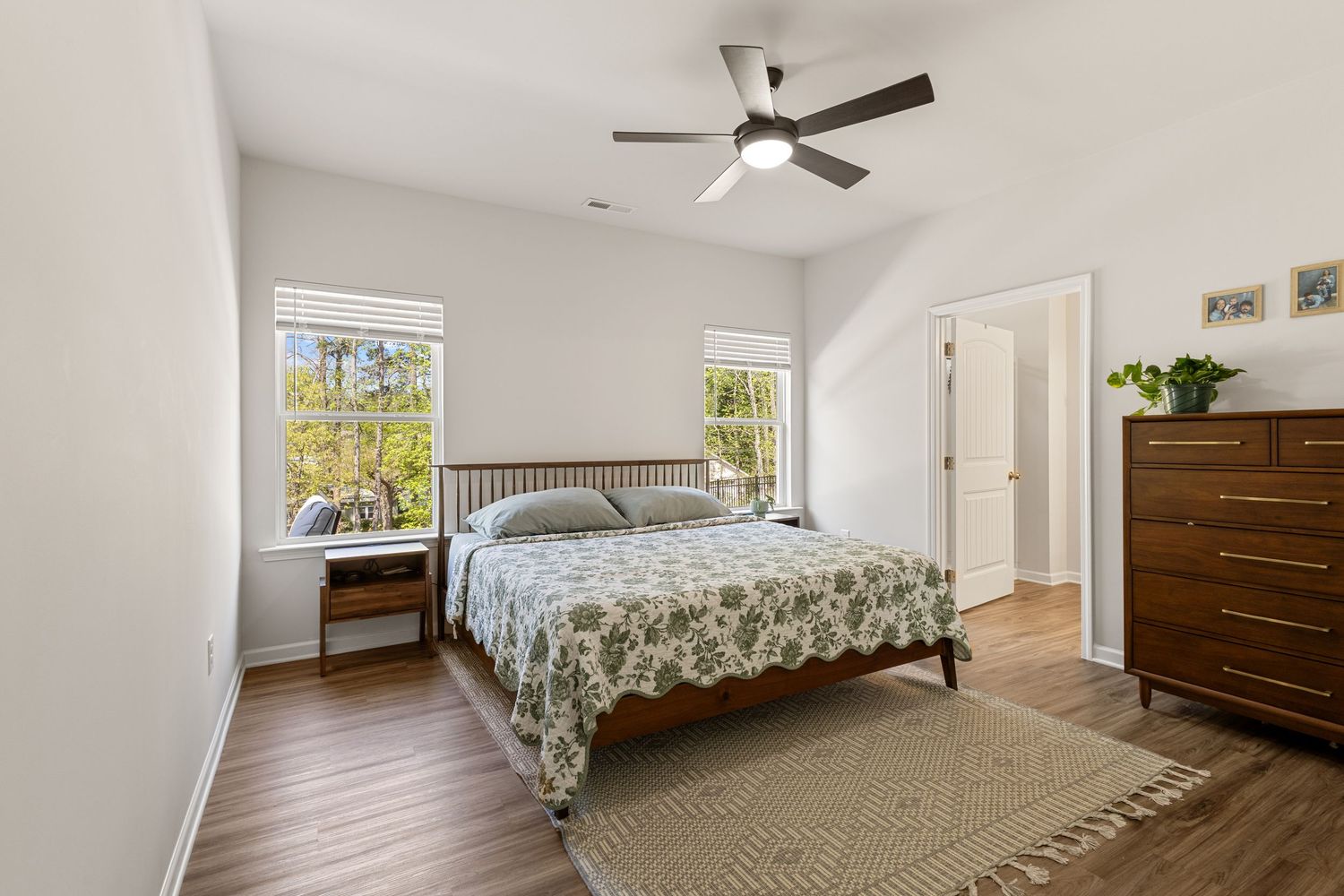 Bright bedroom with hardwood floors, ceiling fan, floral bedspread, wooden dresser, and natural light from double windows.