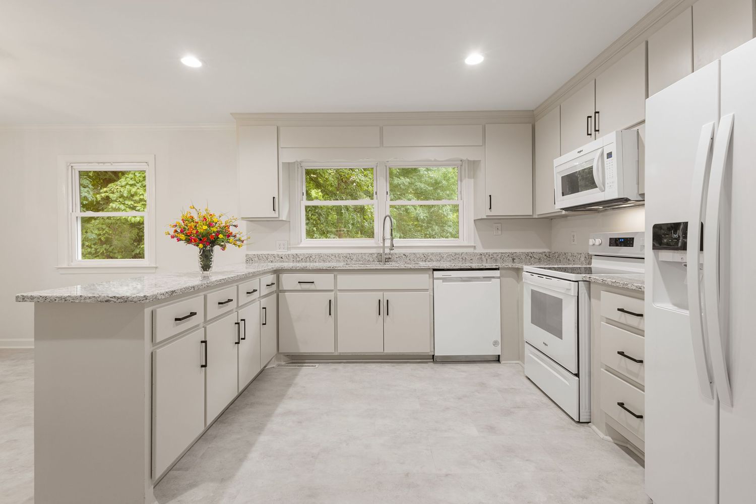 Bright modern kitchen with white cabinets, granite countertops, white appliances, and large windows overlooking greenery.
