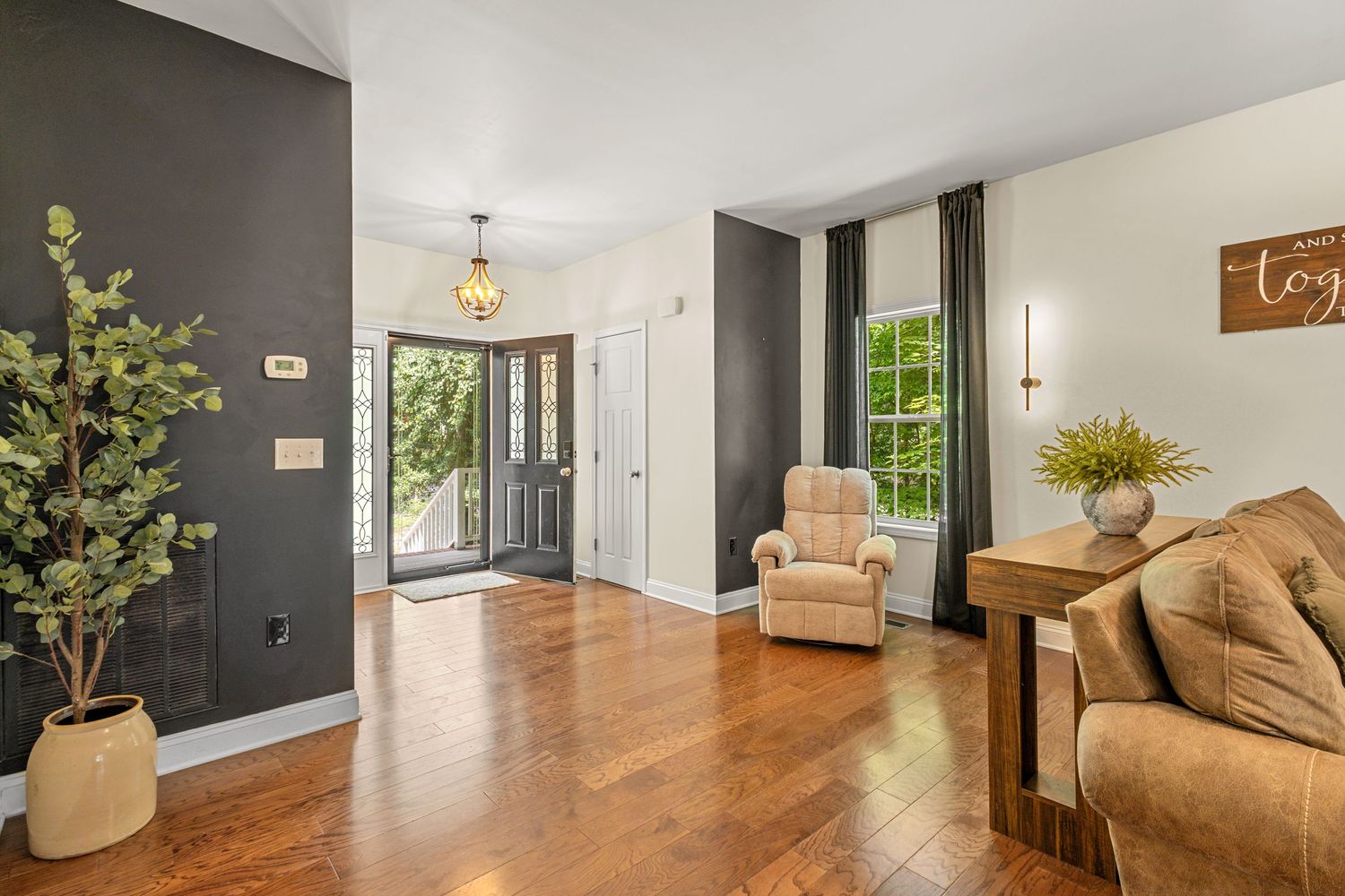 Bright living room with hardwood floors, dark accent walls, beige armchair, and French doors leading to a sunlit entryway.