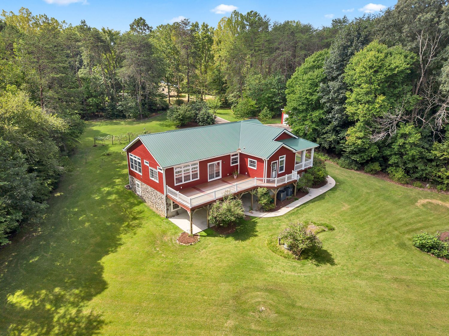 Aerial view of a red farmhouse with green metal roof surrounded by lush trees and manicured lawn in a rural setting.