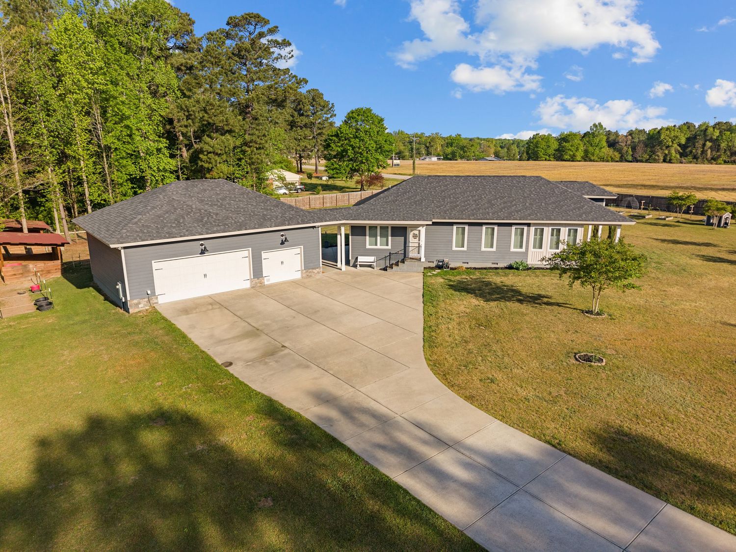 Aerial view of a single-story ranch home with two-car garage, long driveway, and lush green lawn surrounded by trees.