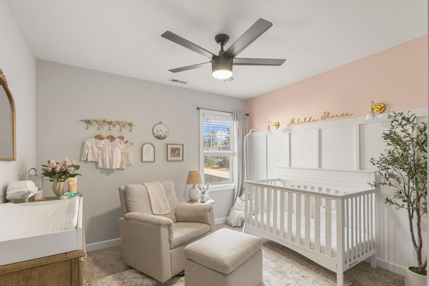 Modern white and beige nursery with ceiling fan features cozy seating, white crib and natural decor elements.