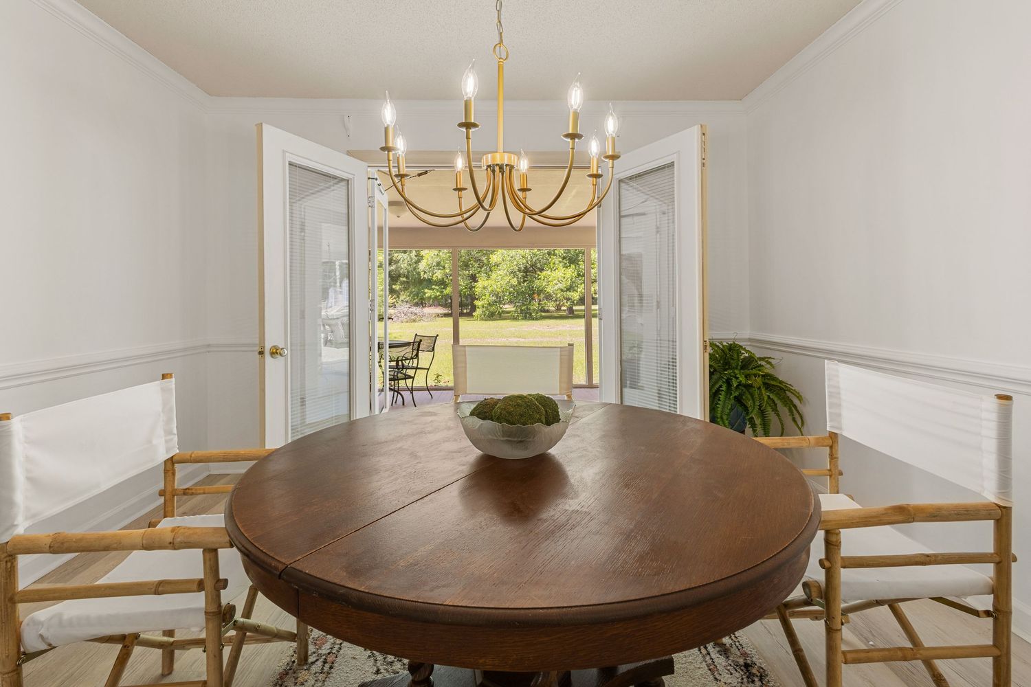Elegant dining room with round wood table, gold chandelier, wainscoting, and French doors opening to a green outdoor view.