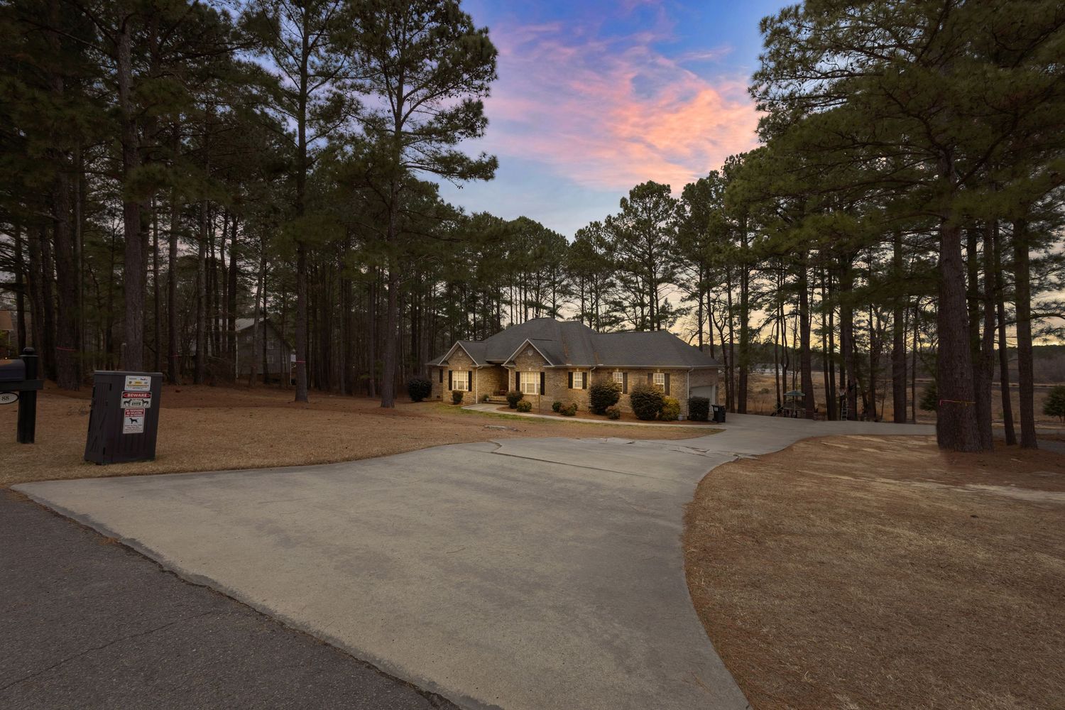 Ranch-style home surrounded by tall pine trees with a curved driveway at dusk under a colorful sunset sky.