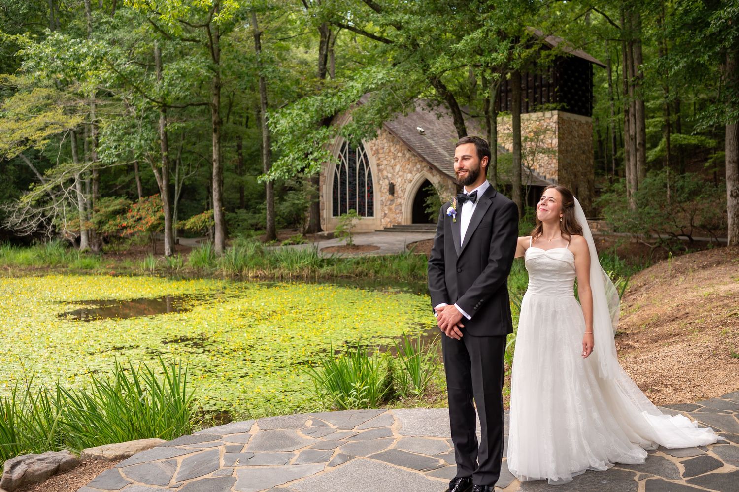 A wedding couple, bride and groom, walks together on a scenic woodland path surrounded by lush greenery.