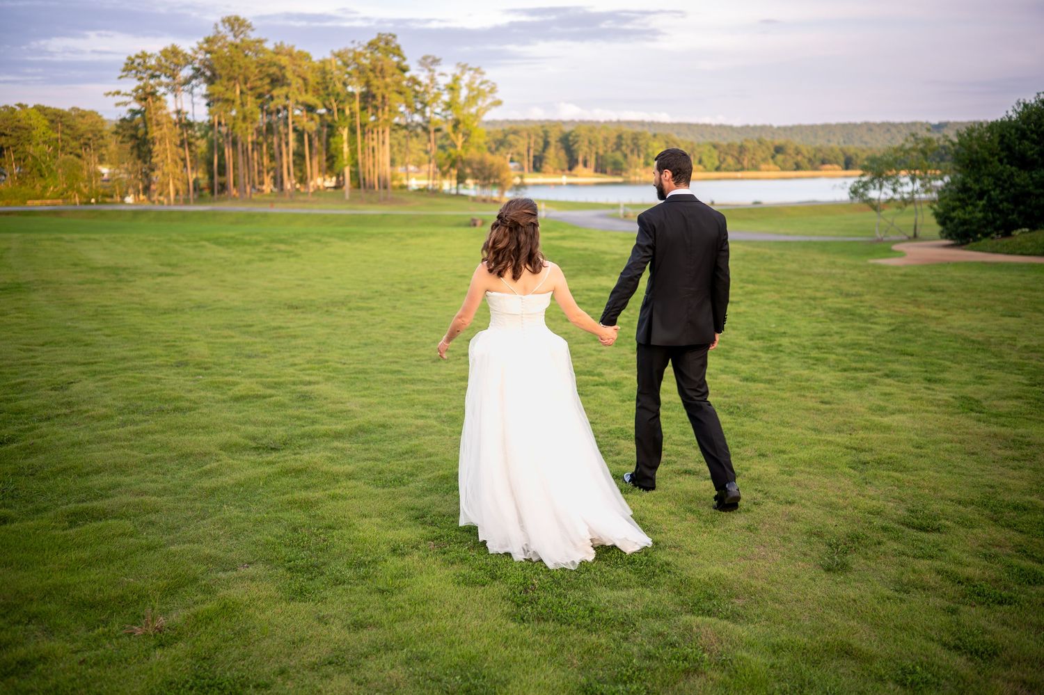 Two people walk hand in hand across a lush green field on a sunny autumn day with trees in the distance.