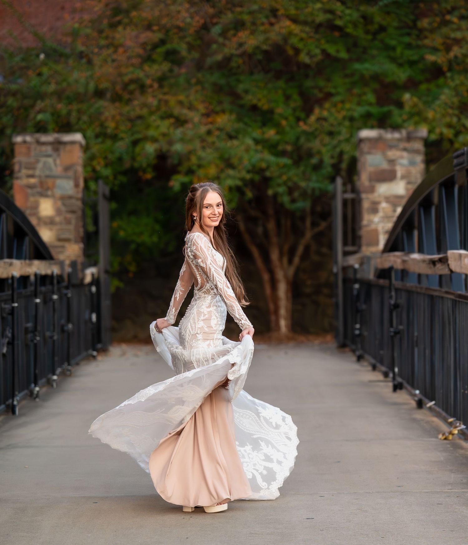Bride in lace gown twirls on stone bridge pathway surrounded by autumn foliage.