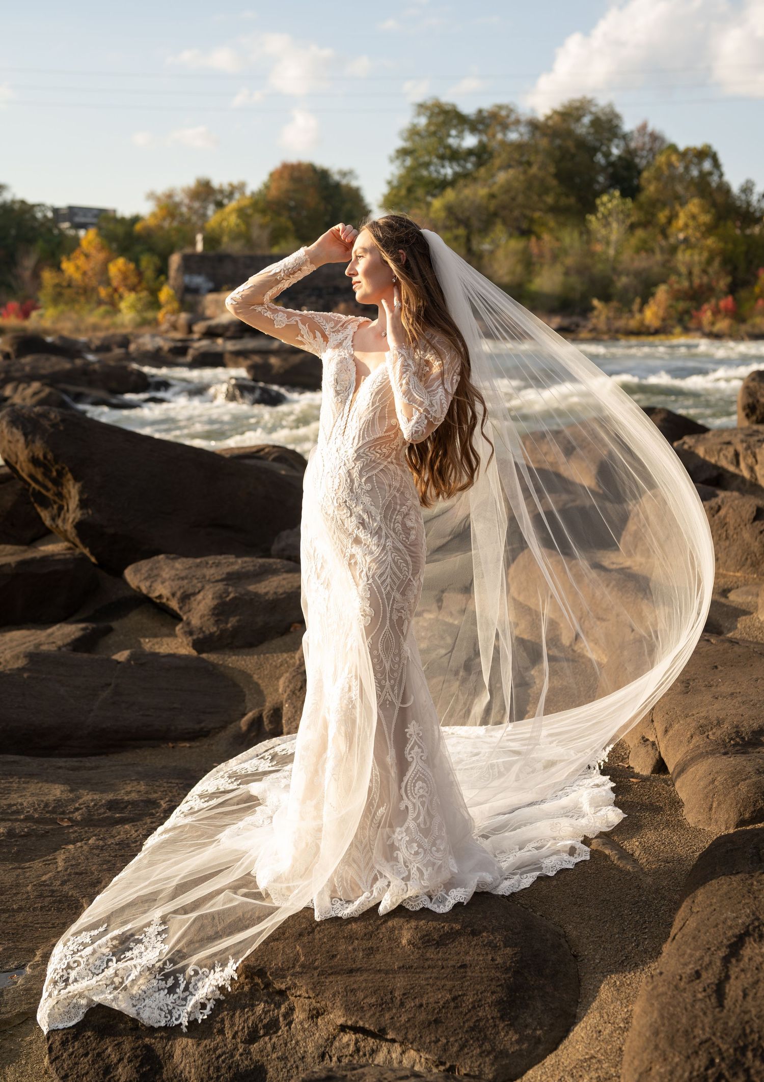 A flowing white wedding dress captured on dark rocks near water during sunset.