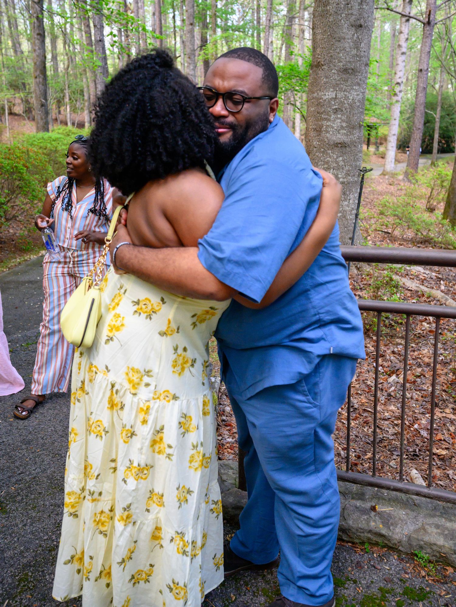 A man in blue scrubs shares a warm embrace with a woman in a yellow floral dress outdoors.
