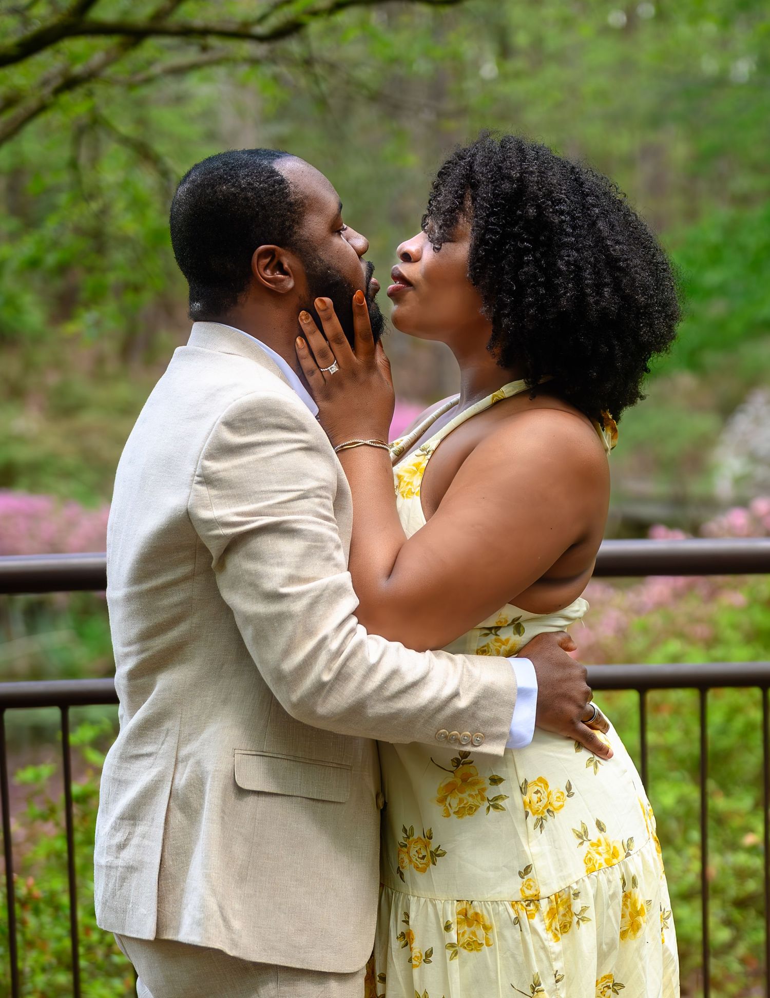Couple shares a romantic kiss near a garden railing surrounded by lush greenery and pink flowers.