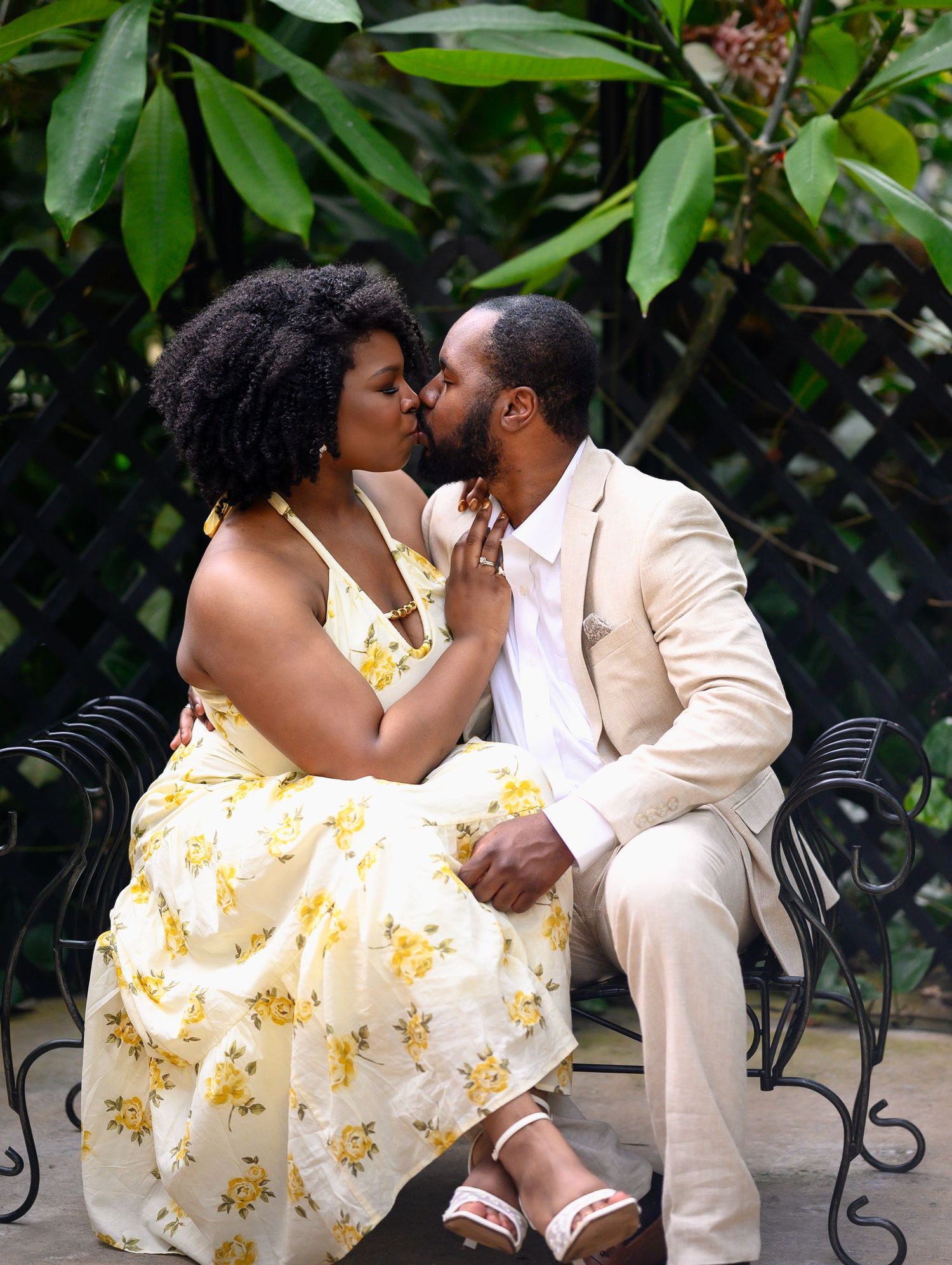 Couple shares a tender kiss while seated on a bench surrounded by tropical greenery.