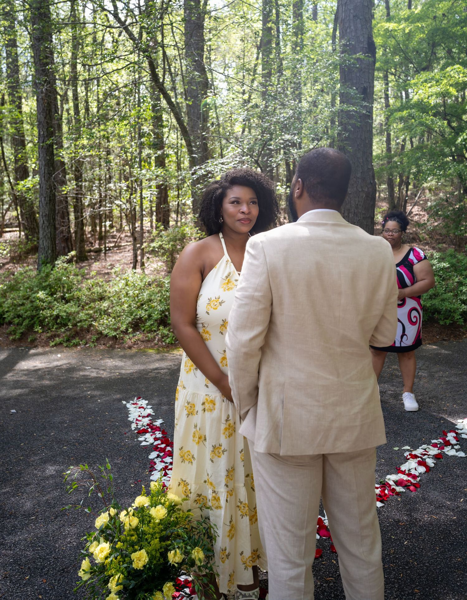 Couple exchanging vows outdoors in a wooded ceremony, woman in yellow floral dress, man in beige suit.