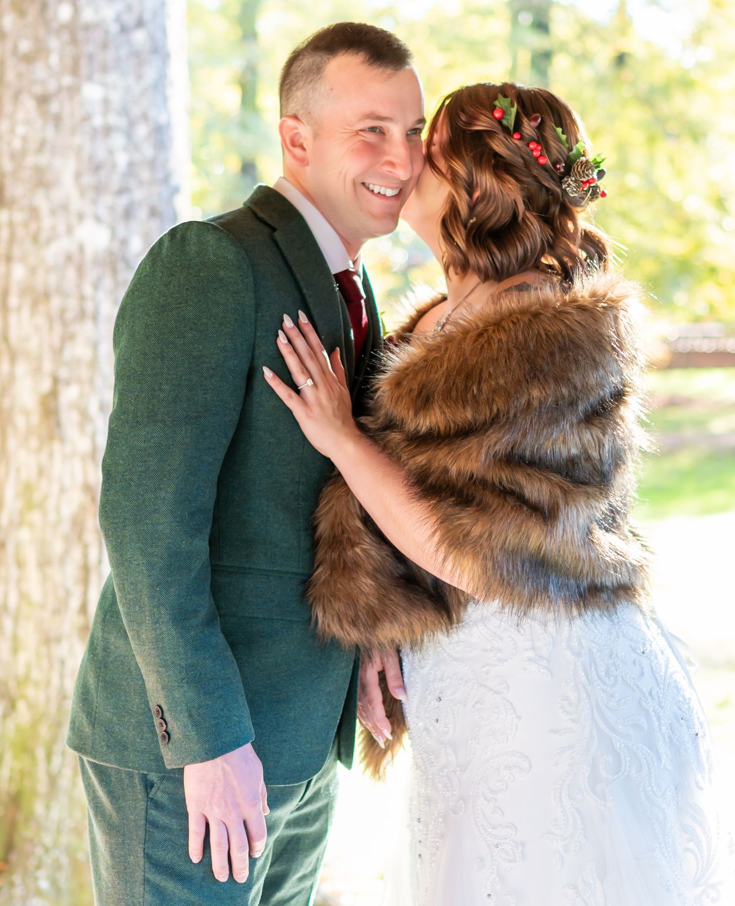 Couple shares romantic moment outdoors with bride wearing fur wrap and green suited groom against natural backdrop.